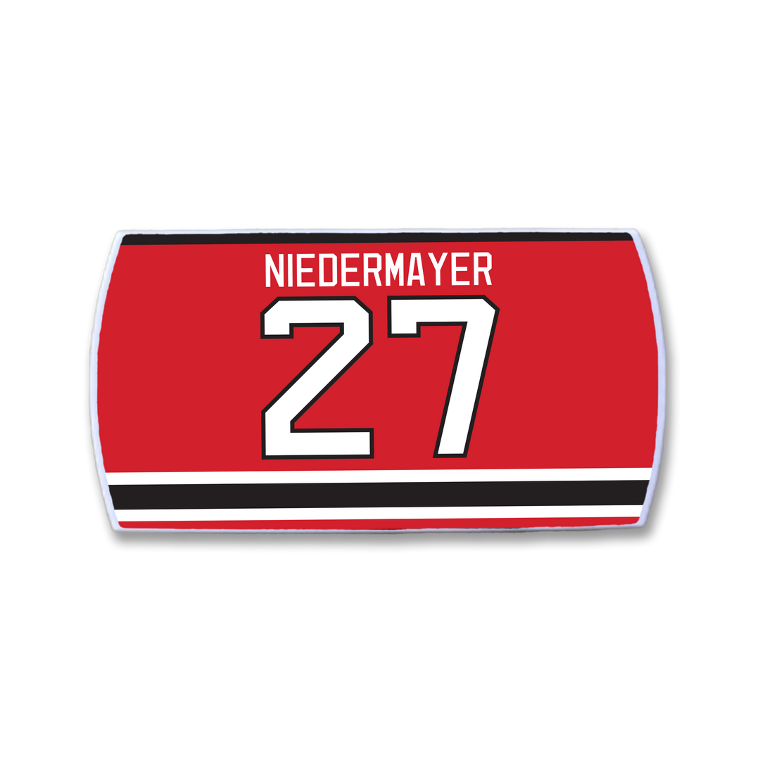 The ZippyBack™ Scott Niedermayer red hockey jersey nameplate displays "Niedermayer" in white above a bold white 27, accented by black and white stripes—celebrating the legacy of leadership and excellence in two-way play.