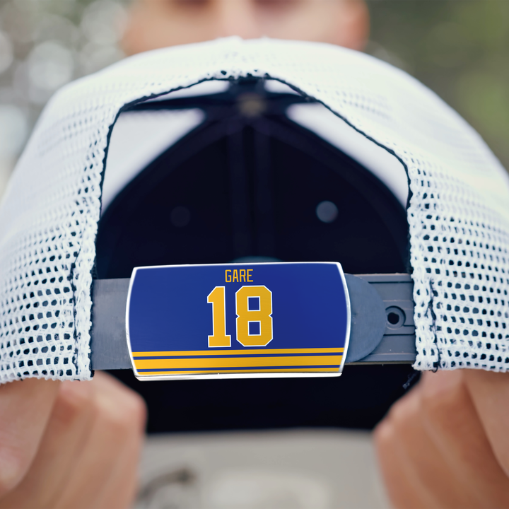 Close-up of a person holding a white mesh ZippyBack™ baseball cap backwards, showing a blue label with yellow stripes, “Danny Gare,” and the number 18 on the adjustable strap—a nod to the hockey scorer’s instinct.