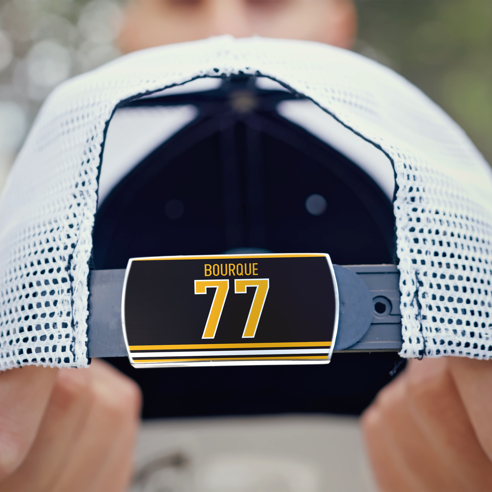 Close-up of a person holding a white mesh ZippyBack™ baseball cap called "Ray Bourque," featuring an adjustable strap with a label reading BOURQUE 77 in yellow and white. The person's face is blurred in the background.