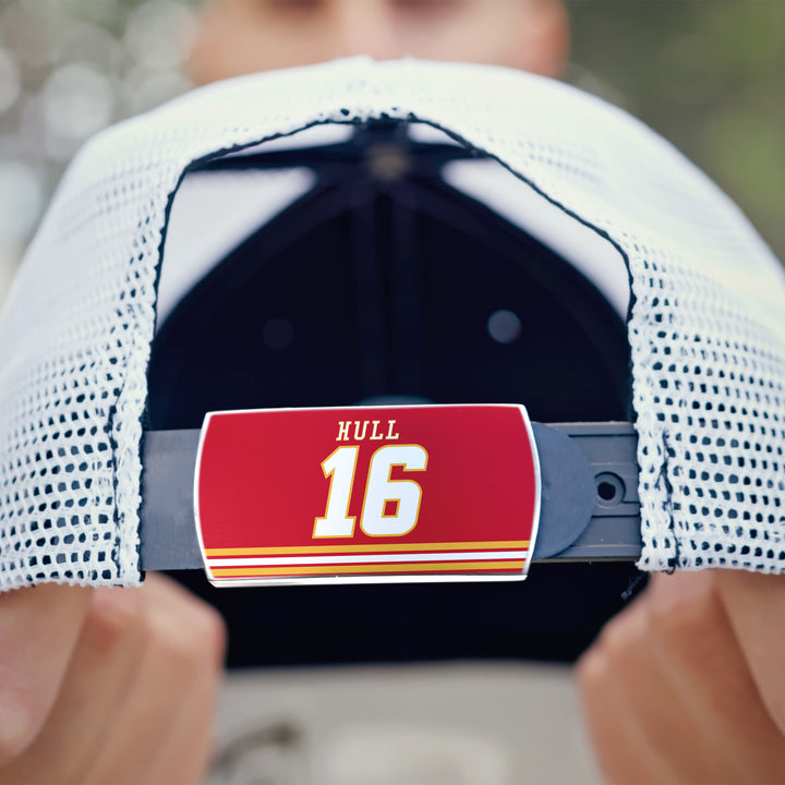 A person holds a white mesh ZippyBack™ baseball cap featuring an adjustable strap with a red label reading "Brett Hull," honoring the legendary hockey goal scorer.