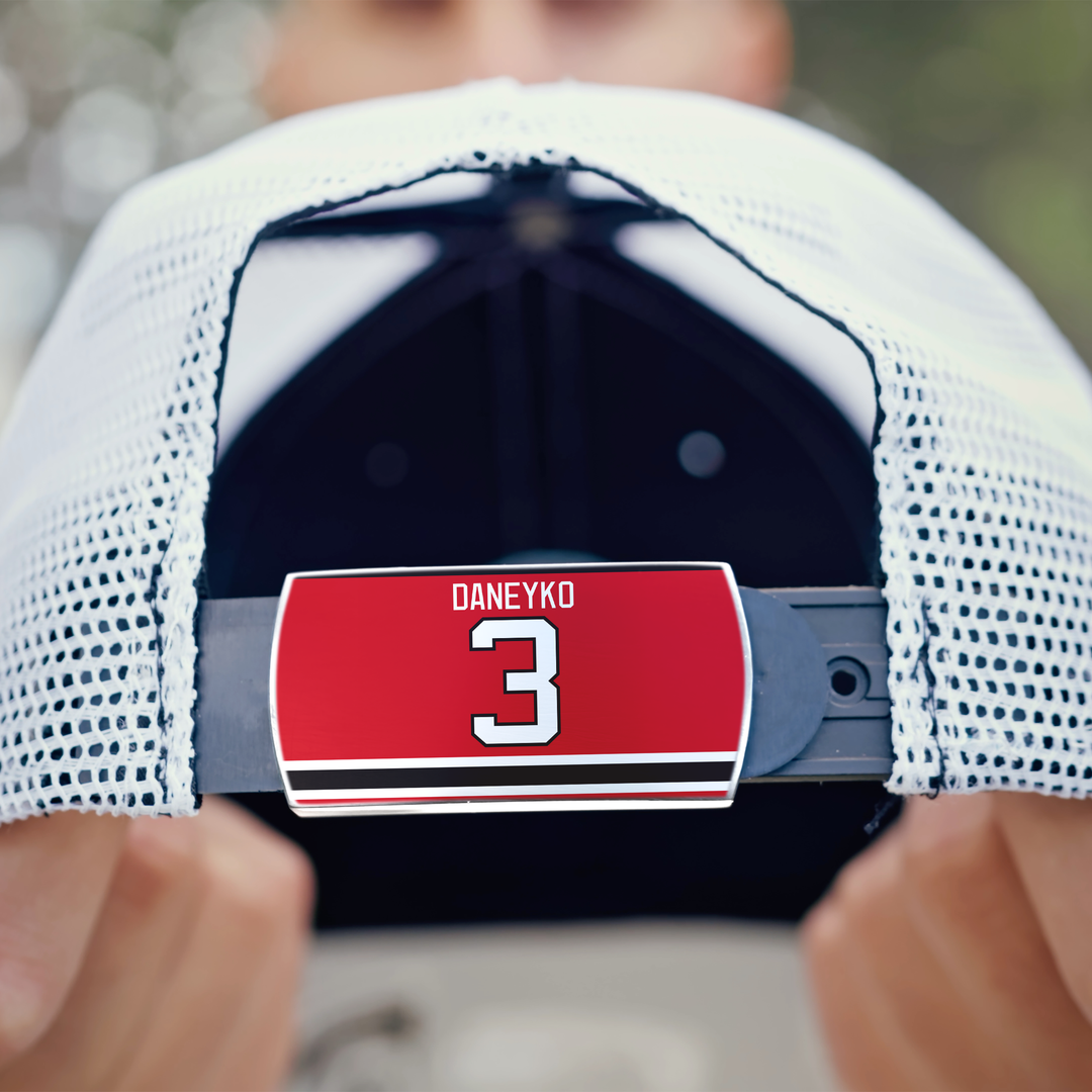 A person holds a white mesh ZippyBack™ Ken Daneyko baseball cap, highlighting the adjustable strap with a red label featuring his name, a bold number 3, and two black stripes—honoring his legendary defense and durability.