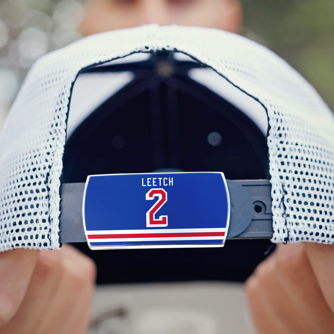 Close-up of the back of a white mesh ZippyBack™ baseball cap. A hand holds the hat, showing a blue LEETCH 2 label on the adjustable strap with stripes underneath, honoring legendary hockey defenseman Brian Leetch.