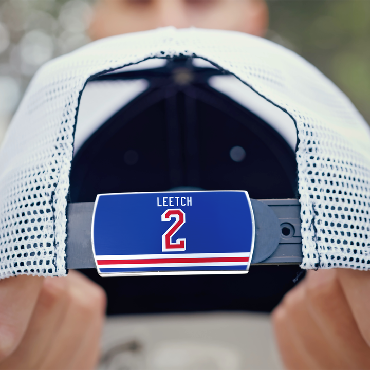 Close-up of the back of a white mesh ZippyBack™ baseball cap. A hand holds the hat, showing a blue LEETCH 2 label on the adjustable strap with stripes underneath, honoring legendary hockey defenseman Brian Leetch.