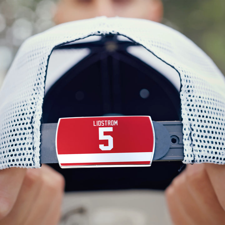 Close-up of a ZippyBack™ white mesh baseball cap featuring a red Nicklas Lidström label with the number 5 on the adjustable strap. Hands hold the cap; a blurred face is visible in the background.