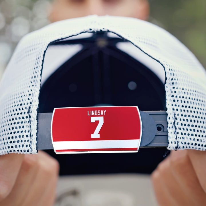 A close-up shows the back of a white mesh ZippyBack™ cap, featuring a red “Ted Lindsay” nameplate above a bold white number 7. Hands hold the hat, paying tribute to hockey legend Ted Lindsay against a softly blurred background.