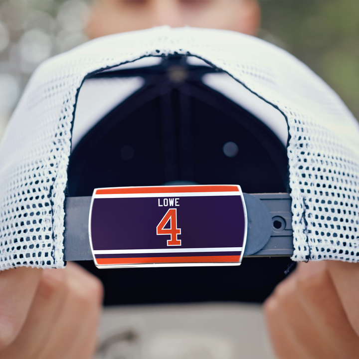 A person holds the back of a white mesh ZippyBack™ Kevin Lowe cap, featuring a "LOWE 4" label in white and orange on a dark blue background with orange and white stripes, honoring Kevin Lowe’s defensive skills.