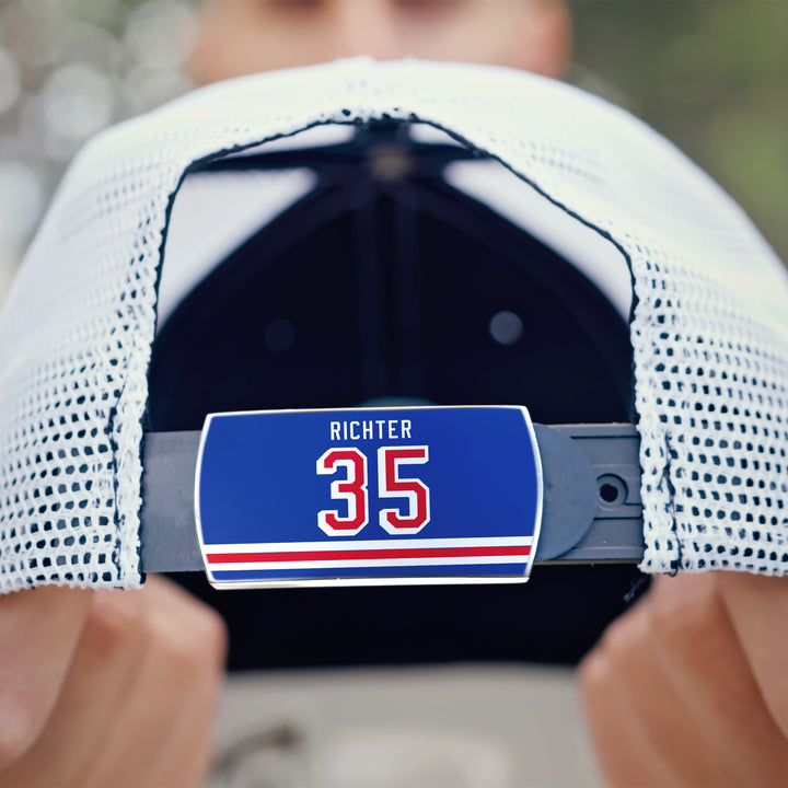 A person holds a white mesh baseball cap with an adjustable strap featuring a blue ZippyBack™ label that reads “Mike Richter” in white and red—a tribute to the legendary goaltender’s performances.