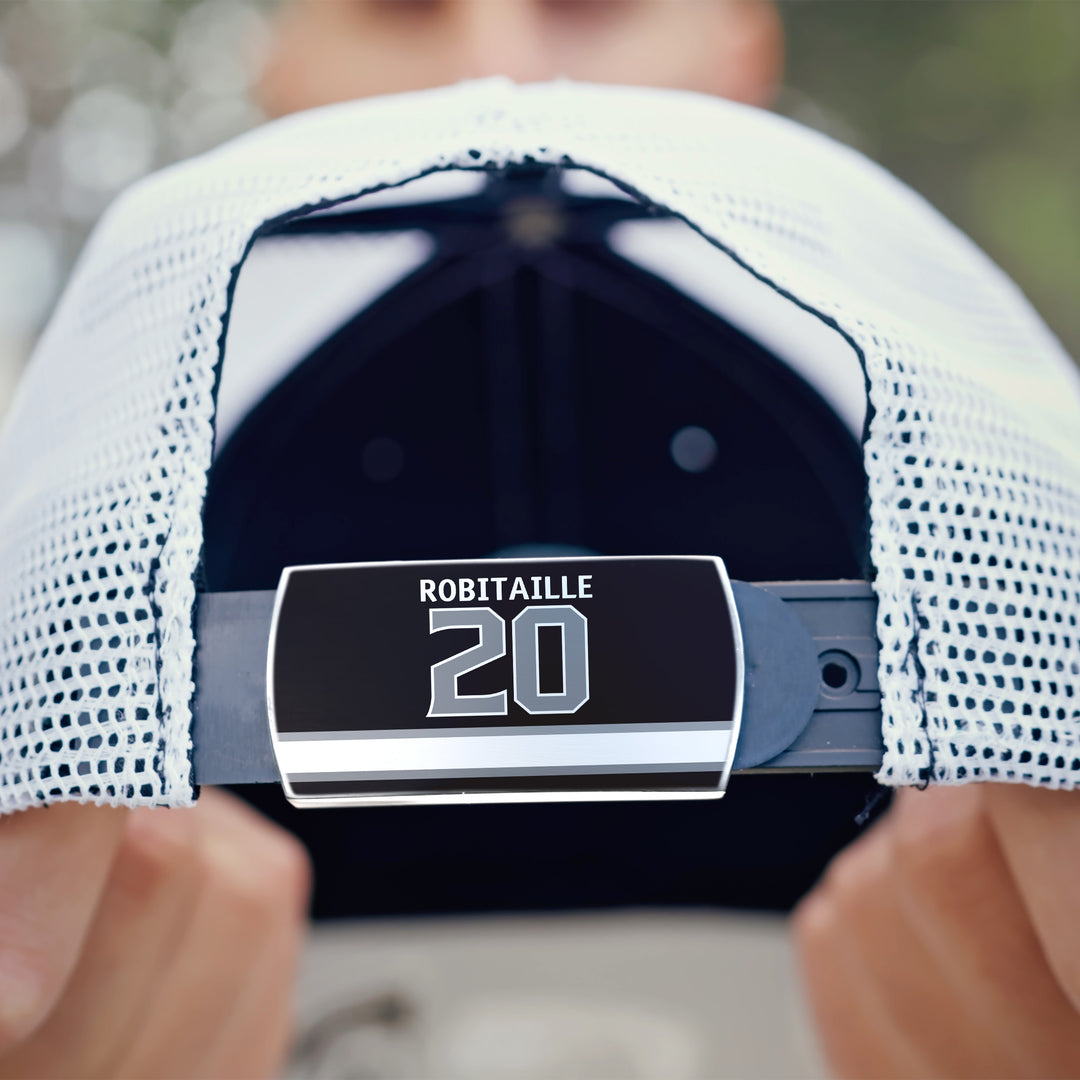 A close-up of someone holding the back strap of a white mesh ZippyBack™ baseball cap with a black and silver Luc Robitaille tag, honoring the legendary hockey left winger.