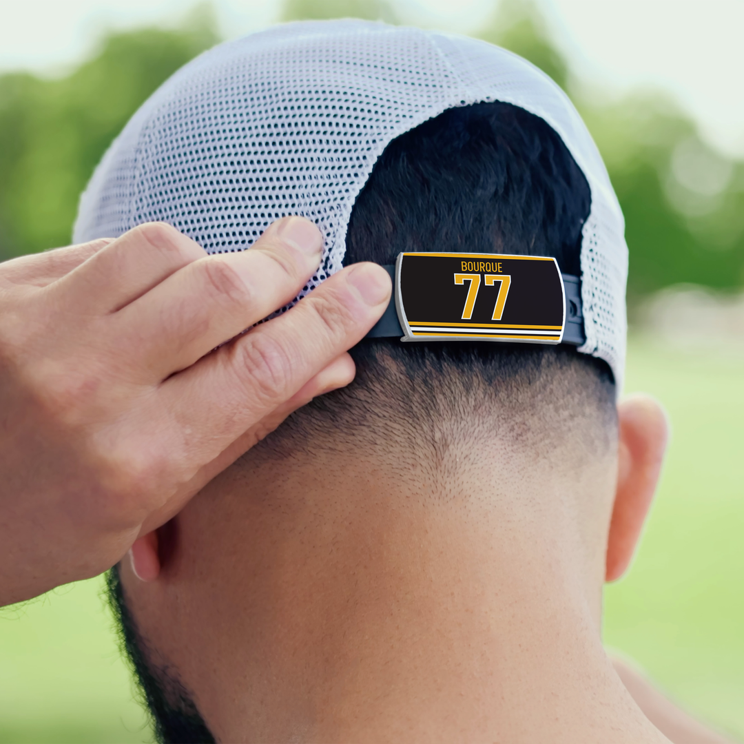 A person wearing a light ZippyBack™ cap adjusts the strap, which features a Ray Bourque tag in black and yellow, with a green park blurred in the background.