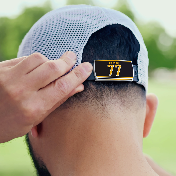 A person wearing a light ZippyBack™ cap adjusts the strap, which features a Ray Bourque tag in black and yellow, with a green park blurred in the background.