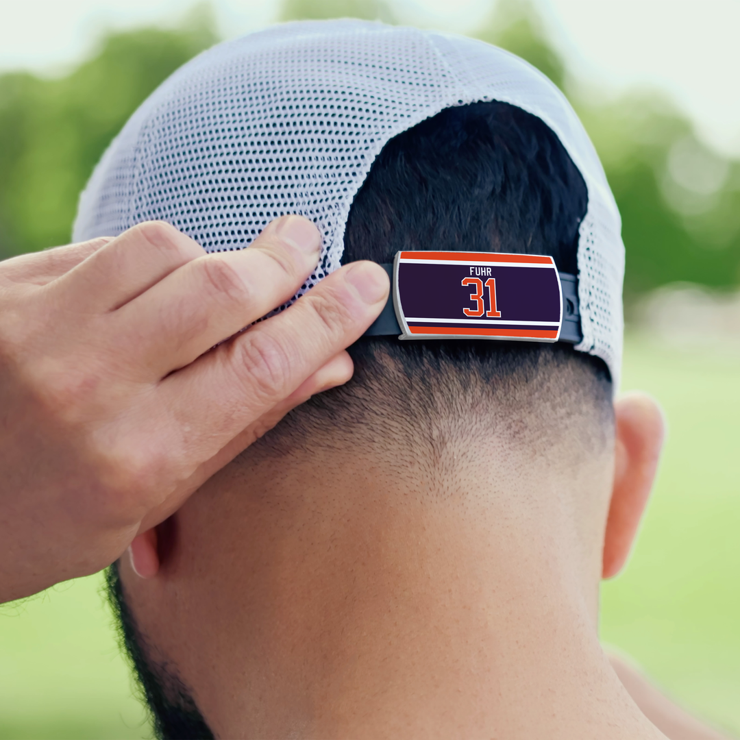 A person adjusts the back of a ZippyBack™ Grant Fuhr mesh baseball cap, featuring a clip with “Fuhr” and “31” in orange and white, against a background of blurred green trees and sunlight.