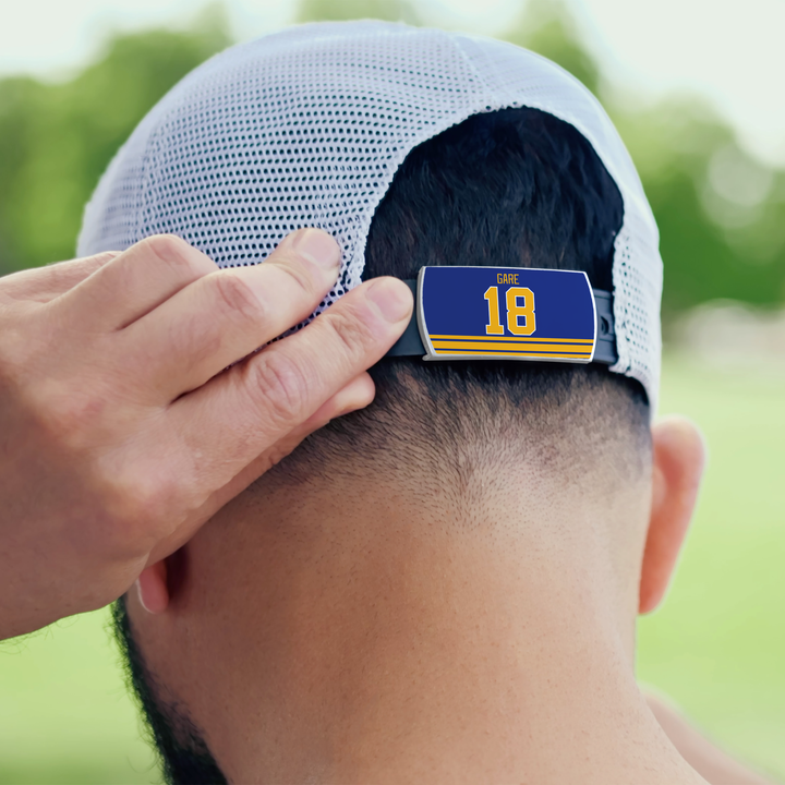 A person adjusts a white mesh ZippyBack™ baseball cap labeled "Danny Gare" in blue and yellow on the back strap, referencing the scorer’s instinct. Greenery appears in the outdoor background.