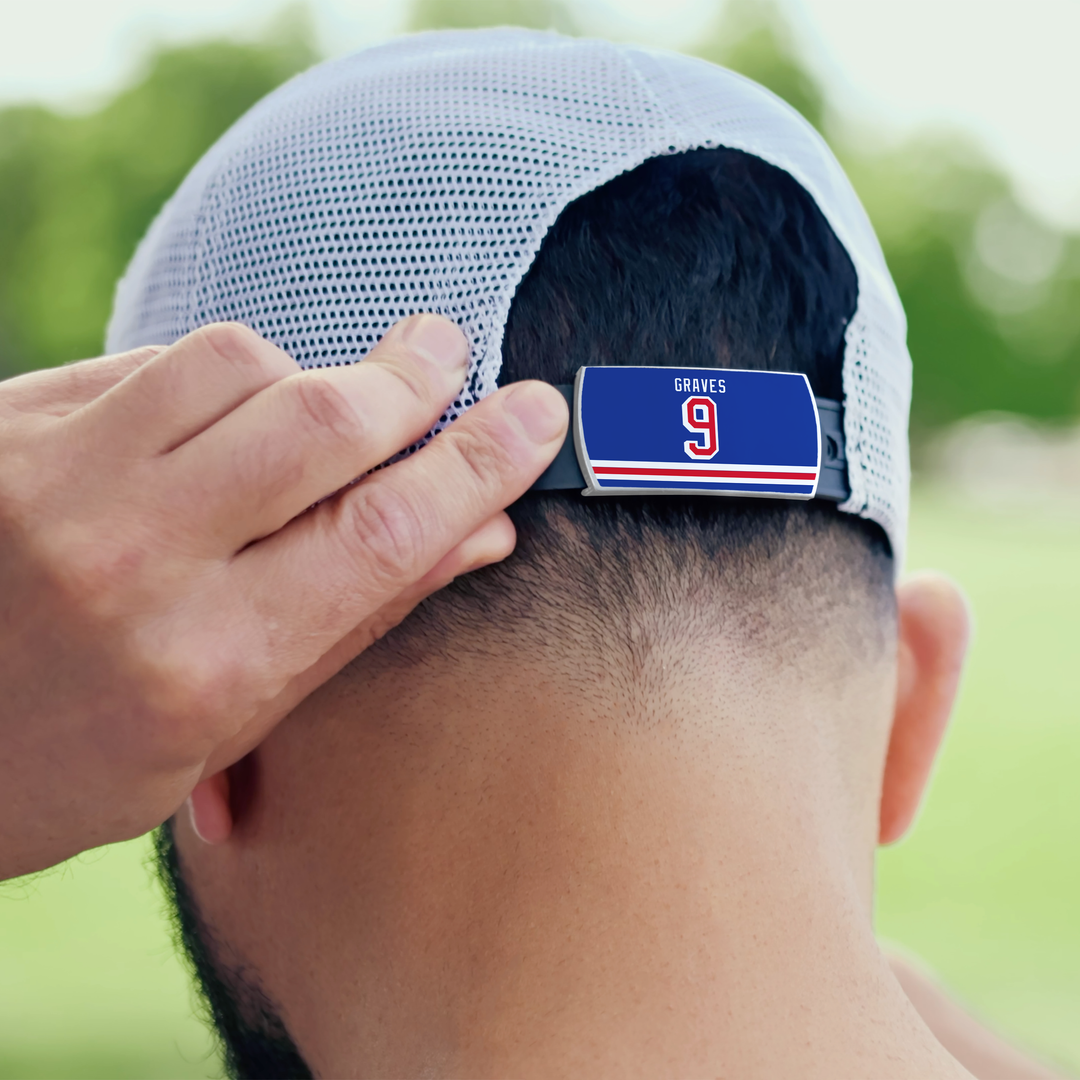 Someone adjusts the back strap of a ZippyBack™ Adam Graves white mesh baseball cap, featuring a blue GRAVES nameplate and red/white number 9. Sunlit green trees blur in the background, capturing true hockey fan energy.