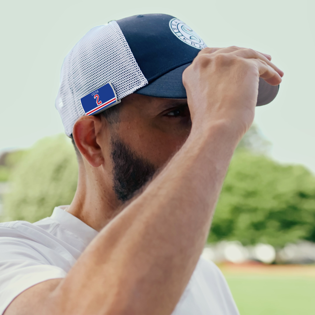 A man with a neatly trimmed beard, hockey defenseman-like, wears a ZippyBack™ Brian Leetch navy and white mesh baseball cap with a blue patch and red "2" on the side. He tilts the brim forward; green foliage blurs in the background.