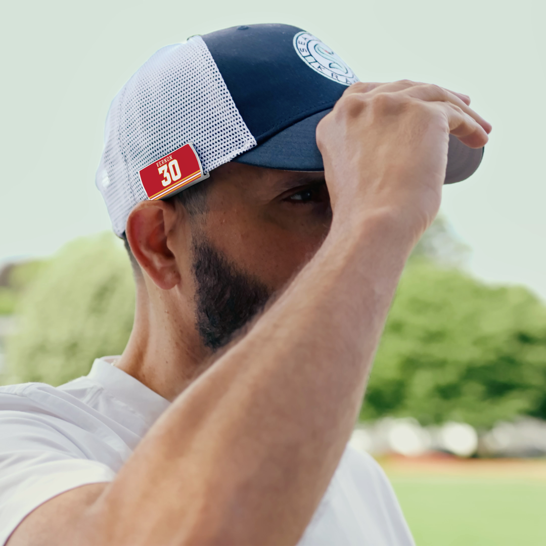A bearded man adjusts his navy and white ZippyBack™ Mike Vernon mesh cap outdoors. A digital red SPF 30 sunscreen label appears on the side. Green trees and a blurred background hint at a sunny day.