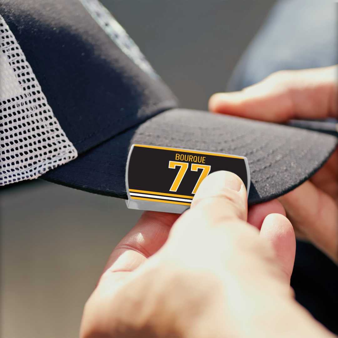 A person attaches a ZippyBack™ Ray Bourque black and yellow sticker to the brim of a black trucker hat with a white mesh back.