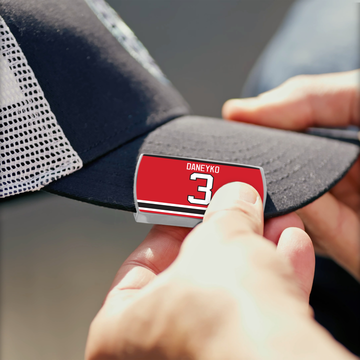 A person attaches a ZippyBack™ Ken Daneyko red and white label reading "DANEYKO 3" to the brim of a black and white mesh cap.