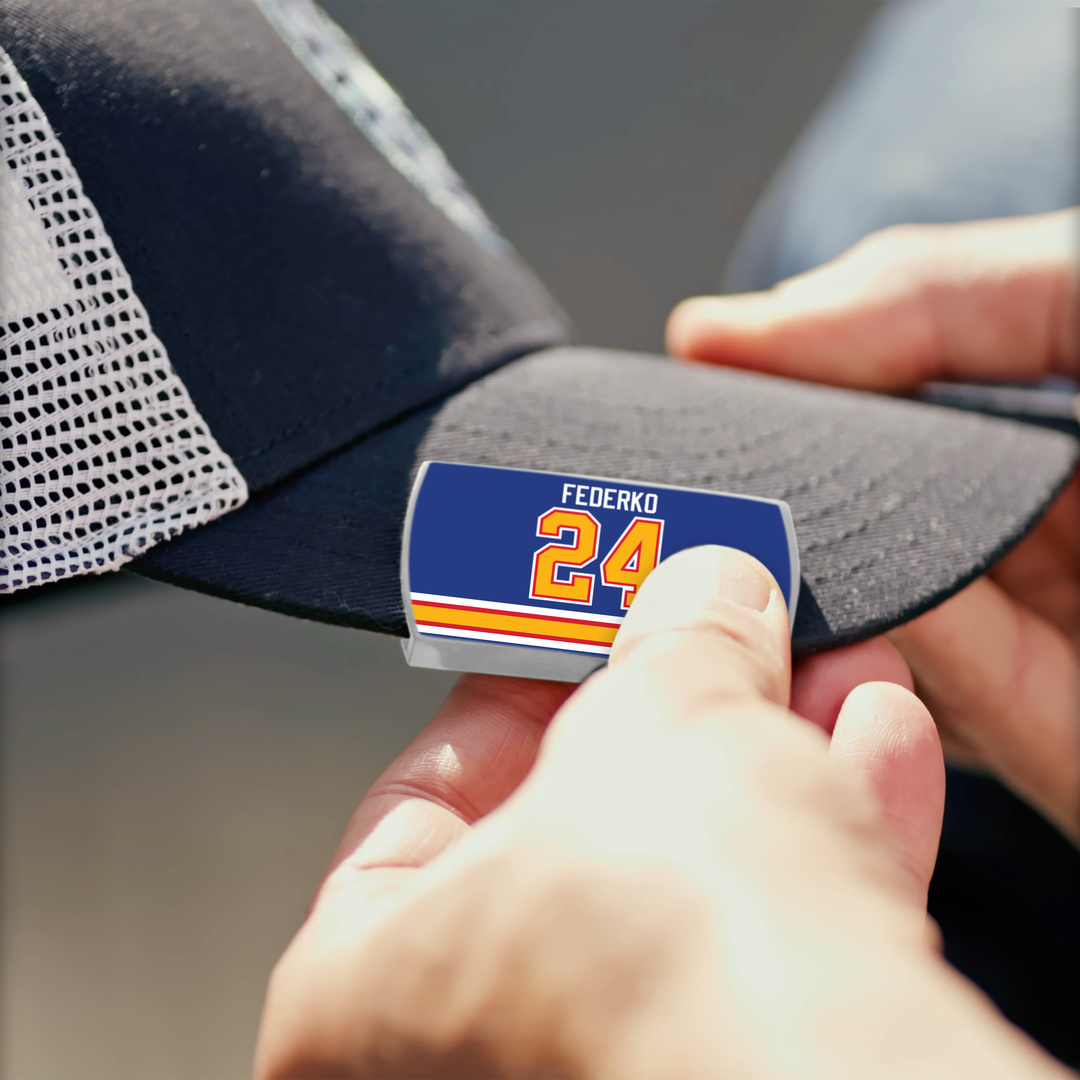A person pins a ZippyBack™ Bernie Federko rectangular pin with blue, white, and orange stripes to the brim of a black mesh baseball cap.