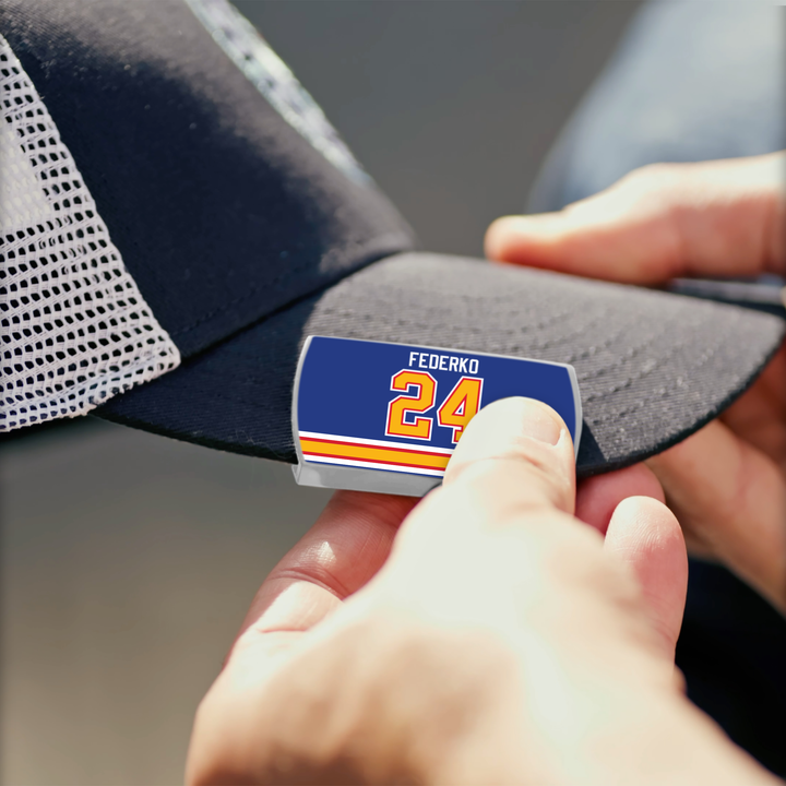 A person pins a ZippyBack™ Bernie Federko rectangular pin with blue, white, and orange stripes to the brim of a black mesh baseball cap.