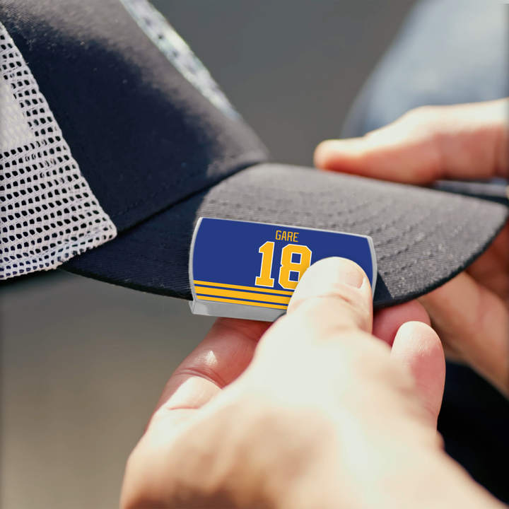A person pins a blue and yellow ZippyBack™ Danny Gare pin to a black mesh baseball cap, honoring his scorer’s instinct.