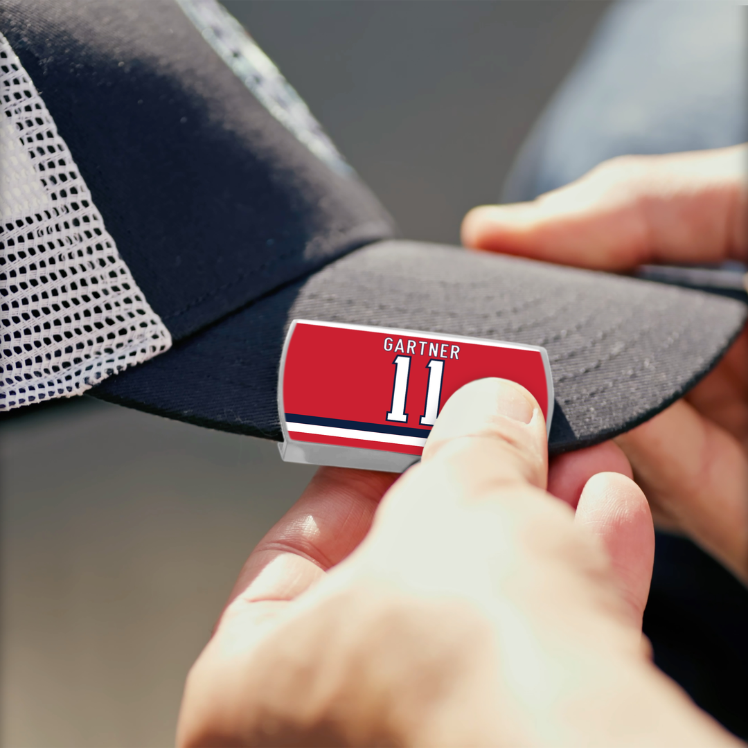 Someone is attaching a red and white ZippyBack™ Mike Gartner name tag sticker reading "Gartner 11" to the black brim of a mesh baseball cap, a nod to Mike Gartner’s legendary speed on the ice.