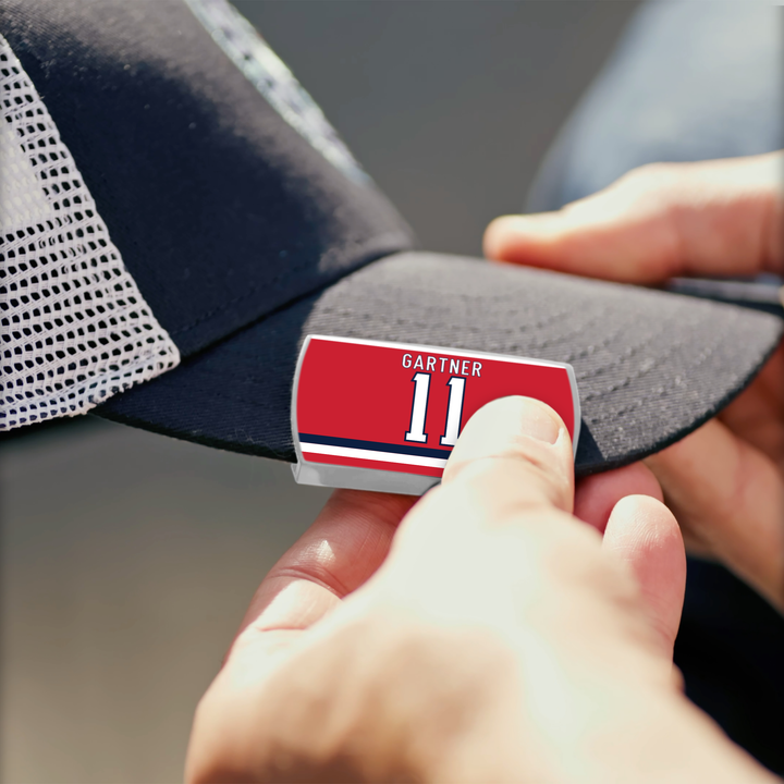 Someone is attaching a red and white ZippyBack™ Mike Gartner name tag sticker reading "Gartner 11" to the black brim of a mesh baseball cap, a nod to Mike Gartner’s legendary speed on the ice.