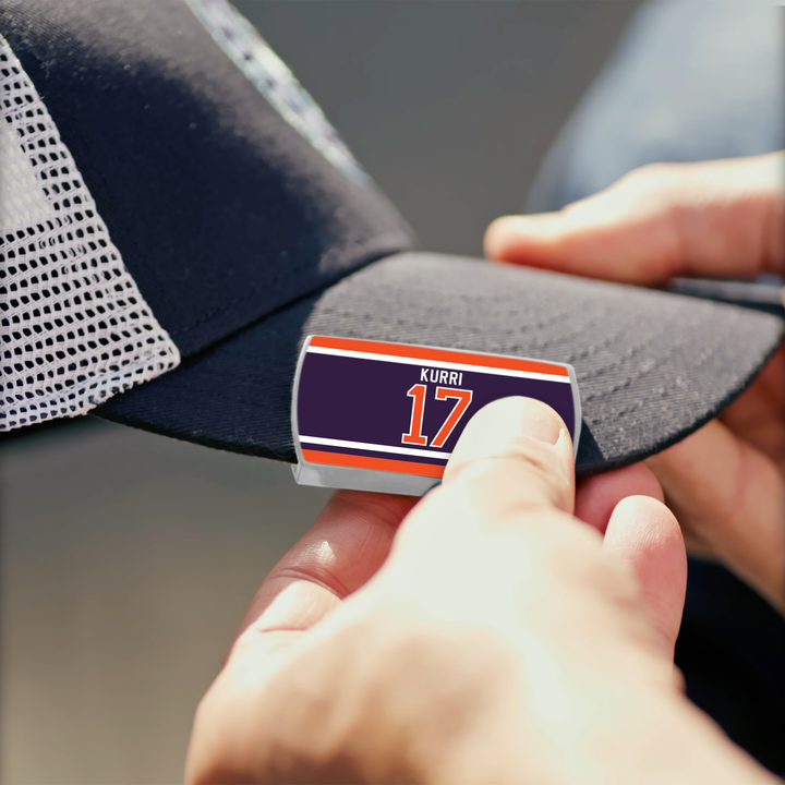 A person adds a ZippyBack™ Jari Kurri sticker, featuring the name and number 17 in orange and navy blue, to the brim of a dark mesh-back baseball cap.