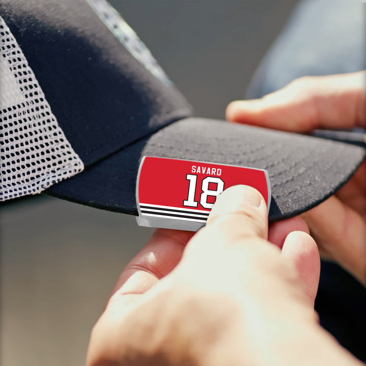 A person attaches a red ZippyBack™ Denis Savard 18 nameplate to a black and white mesh baseball cap, celebrating Denis Savard’s legendary hockey skill and puck handling.