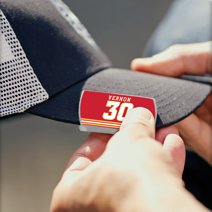 A person swiftly attaches a ZippyBack™ Mike Vernon red and yellow name and number sticker to the bill of a black mesh baseball cap, showcasing speed and precision.