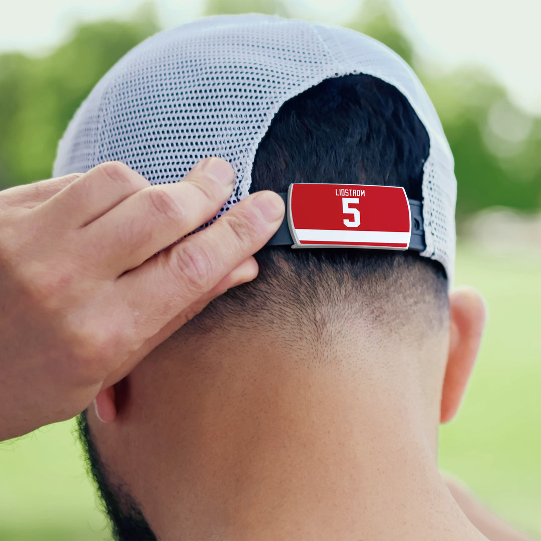 Someone adjusts the back strap of a white mesh ZippyBack™ baseball cap, which displays a red name tag with Nicklas Lidström’s name and number 5. Blurred greenery in the background suggests admiration for the hockey legend.
