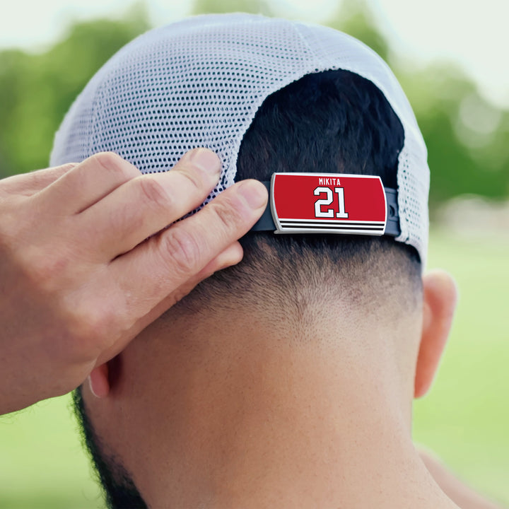 A person adjusts the back strap of a white mesh ZippyBack™ Stan Mikita baseball cap, which displays a red tag honoring hockey legend Stan Mikita and his number 21. Out-of-focus greenery appears in the background.
