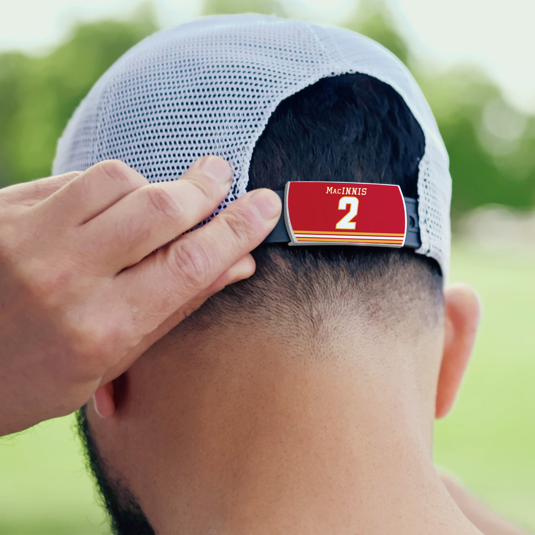 A person adjusts a white mesh ZippyBack™ baseball cap featuring a red name tag with "MacINNIS" and the number 2, honoring legend Al MacInnis. The background is blurred greenery.
