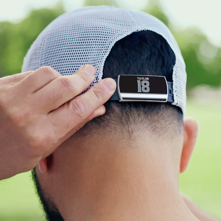 A person adjusts a white mesh ZippyBack™ Dave Taylor cap with a back digital display showing “Taylor” and “18,” honoring playmaker Dave Taylor. Blurred greenery appears in the background.