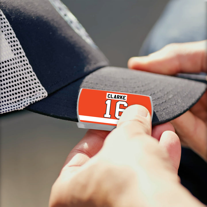 A person attaches a rectangular orange and white ZippyBack™ label reading "Bobby Clarke 16" to a black and white mesh baseball cap, paying tribute to the elite two-way leader.