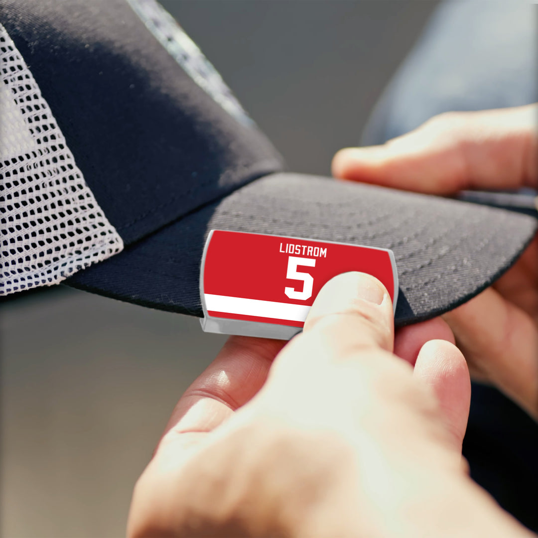 A person attaches a ZippyBack™ Nicklas Lidström badge to the brim of a black and white mesh baseball cap.
