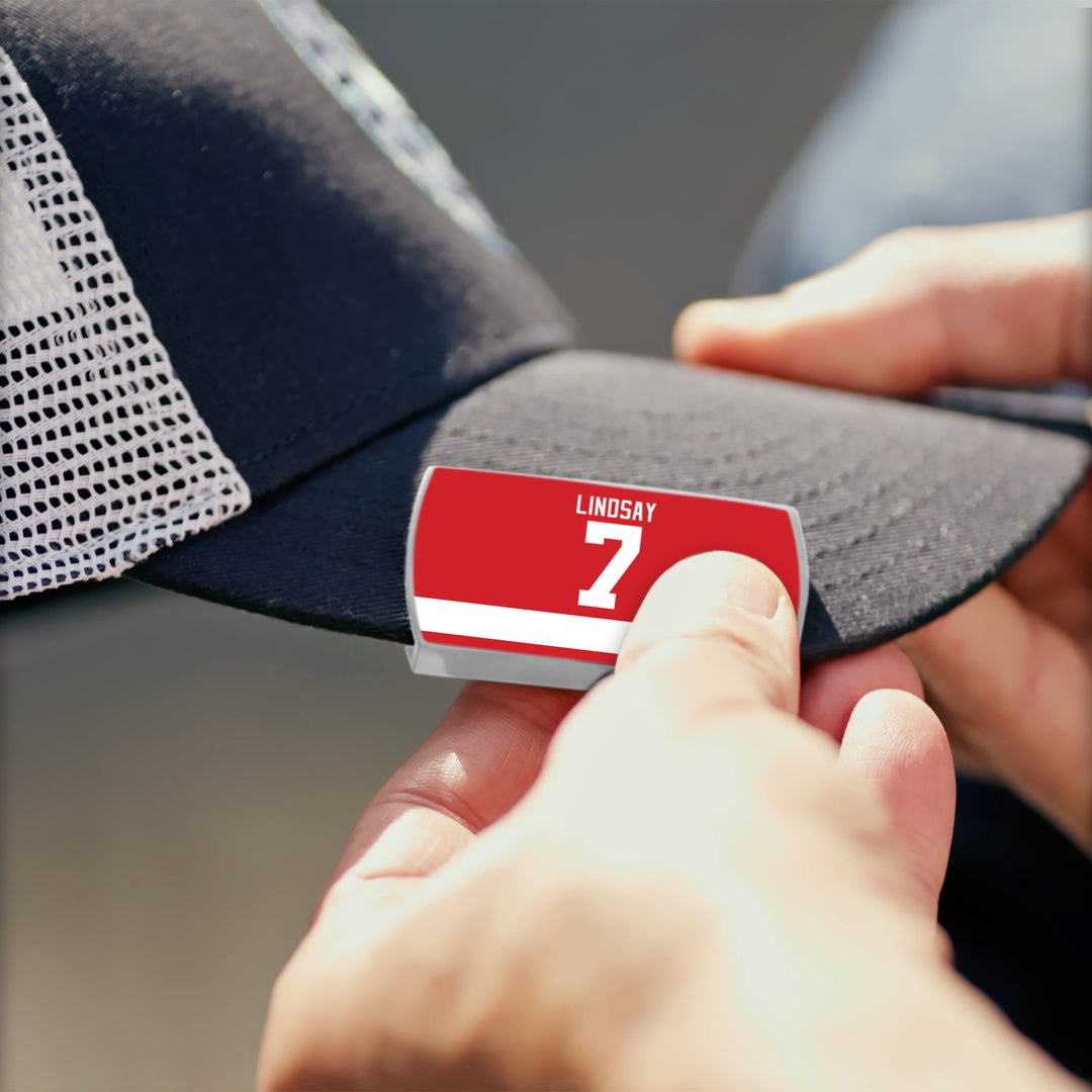 Someone attaches a red ZippyBack™ nameplate with "LINDSAY" and the number 7 to a black mesh cap, honoring hockey legend Ted Lindsay.