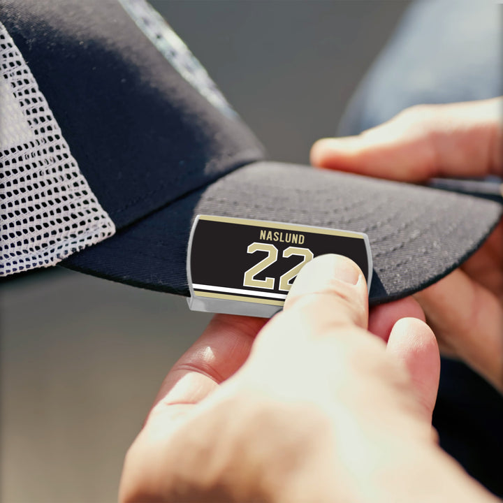 A person fastens a ZippyBack™ Markus Näslund black and gold rectangular pin to a black mesh baseball cap, honoring Näslund&
