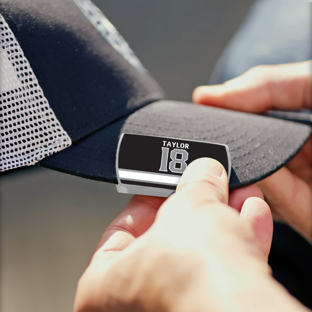 A person attaches a black and silver TAYLOR 18 label to the brim of a black and white ZippyBack™ baseball cap, honoring playmaking forward Dave Taylor&