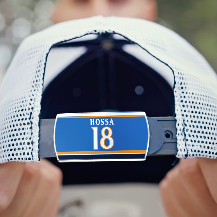 Close-up of a person holding the back of a ZippyBack™ Marian Hossa mesh baseball cap, featuring a blue tag with his name and number 18. The blurred background highlights this tribute to the hockey legend.