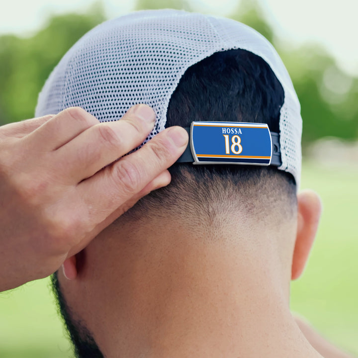 Someone adjusts the back strap of a ZippyBack™ Marian Hossa mesh baseball cap, displaying a blue and gold "Hossa 18" label. Green trees appear blurred in the background.