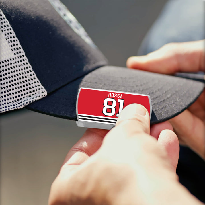 Close-up of hands attaching a red and white ZippyBack™ Marian Hossa pin, featuring the number 81, to the brim of a black and white mesh baseball cap.